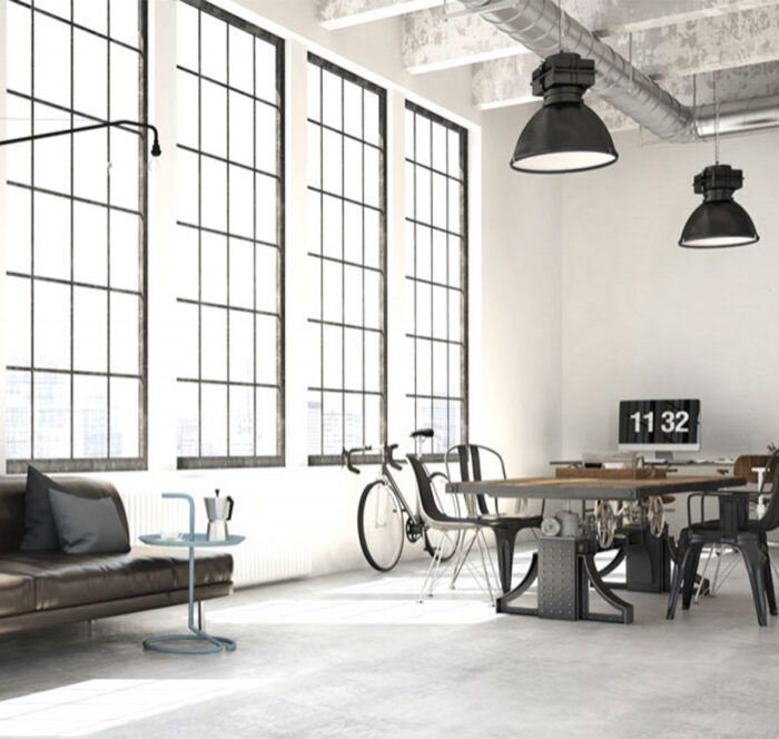 Pièce industrielle style "factory" avec béton brut, mobilier en métal, cuir et bois, plafond haut avec poutres et conduits métalliques visibles, suspensions noires et grande verrière sur tout le mur apportant luminosité.
