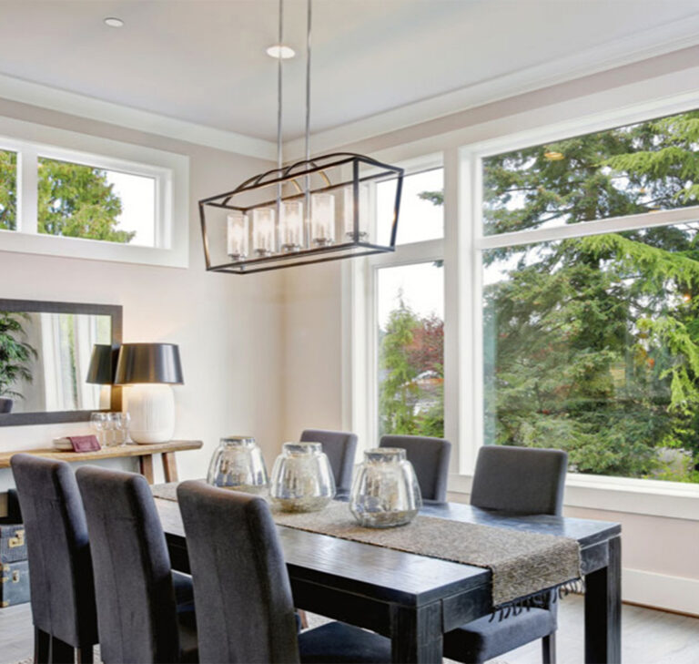 Salle à manger moderne avec une grande table rectangulaire en bois noir massif, placée devant un mur de grandes fenêtres blanches. La pièce est lumineuse, ouverte sur une vue verdoyante extérieure et baignée de lumière naturelle.
