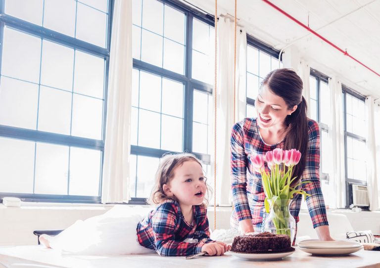 Moment familial dans une pièce à vivre de style industriel, très lumineuse grâce à la lumière naturelle entrant par une grande verrière murale, où une mère souriante regarde sa fille souffler les bougies sur un gâteau