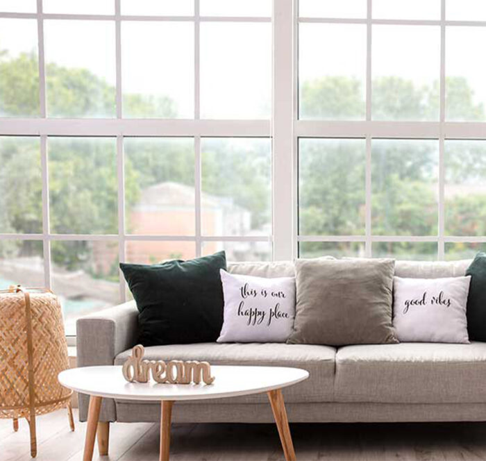 Canapé avec coussins verts, beige et gris dans un salon éclairé par une baie vitrée en menuiserie blanche à croisillons