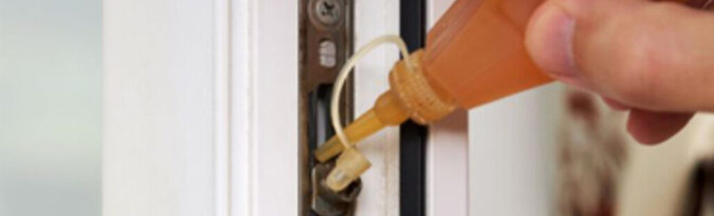 Entretien de fenêtre : une main masculine applique de l’huile sur les mécanismes d’une fenêtre, notamment les ferrures, charnières et poignée, pour tester leur fonctionnement et garantir l’étanchéité de la fenêtre.